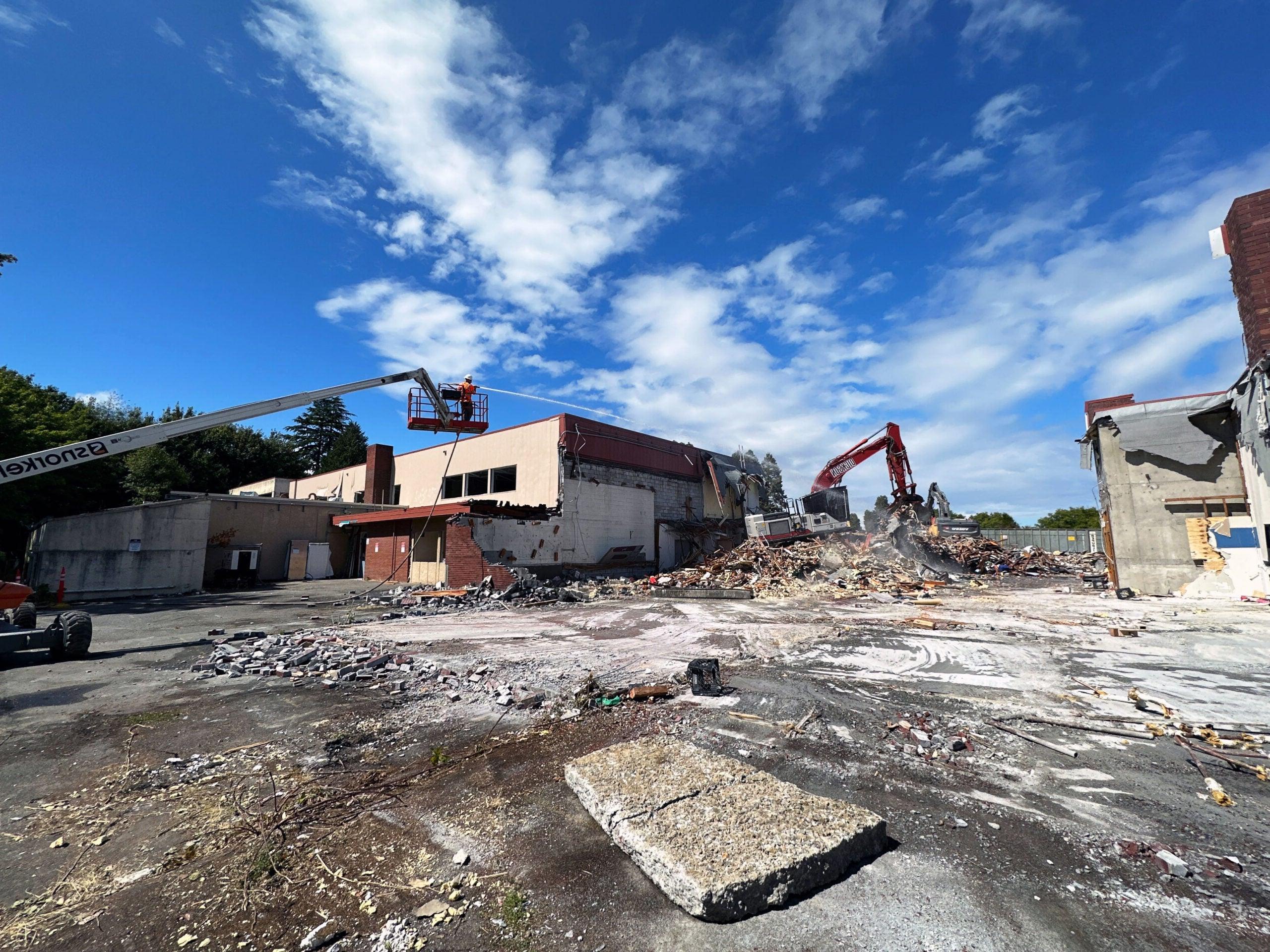 an excavator is crushing a brick building. There is a person on a lift spraying a hose. There is rubble on the concrete in the foreground